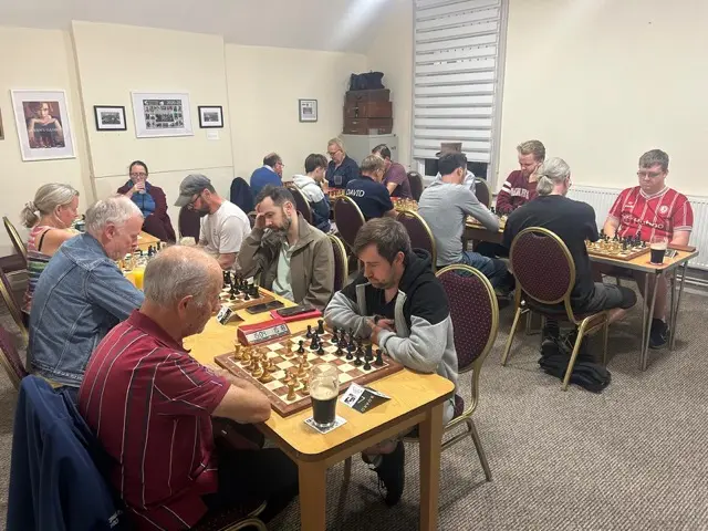 Wide view of the playing room with every board in use, the Queen's Gambit poster on the wall behind